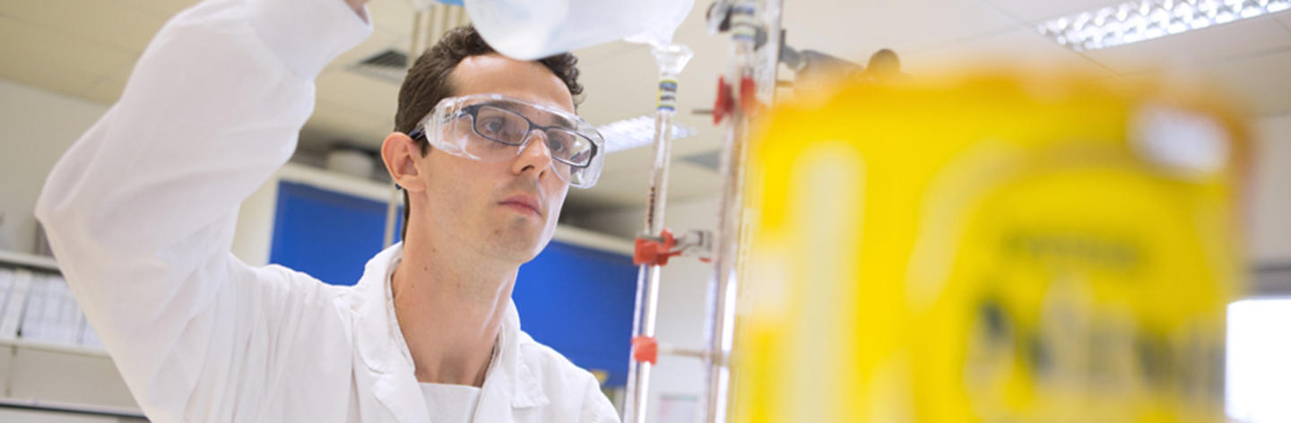 Homem de jaleco branco e óculos de proteção despeja um líquido em um tubo de laboratório.