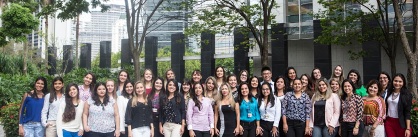 Grupo de mais de 30 mulheres posa para a foto em frente a uma empresa. Algumas usam roupa social e algumas usam crachás. 